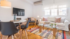 Een moderne woonkamer met een ronde glazen salontafel, een witte bank met gekleurde kussens, een rieten fauteuil, en een eetgedeelte met een glazen tafel en stoelen.