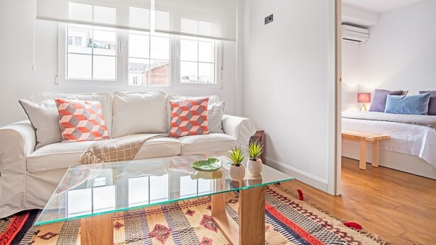 Een moderne woonkamer met een glazen salontafel, een witte bank met decoratieve kussens, een groot raam en uitzicht op het omliggende landschap.