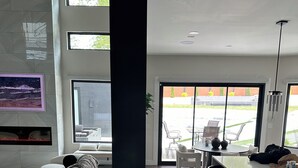 A modern living room with a large sectional sofa, a flat-screen TV mounted on the wall, and a dining area with a long table and bar stools. - Gorgeous Modern Compound w/ private resort backyard & full outdoor kitchen (Decatur)