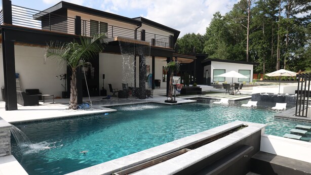 A modern outdoor pool area with a waterfall feature, a seating area with umbrellas, and a building with a balcony. - Gorgeous Modern Compound w/ private resort backyard & full outdoor kitchen (Decatur)