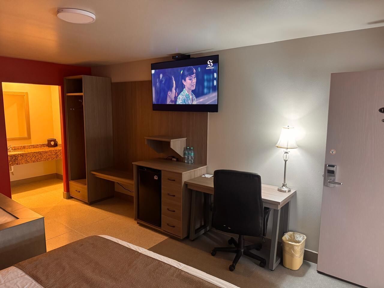 Une chambre d’hôtel comprenant un lit, un bureau avec une chaise, une télévision, une lampe, une poubelle et une armoire.