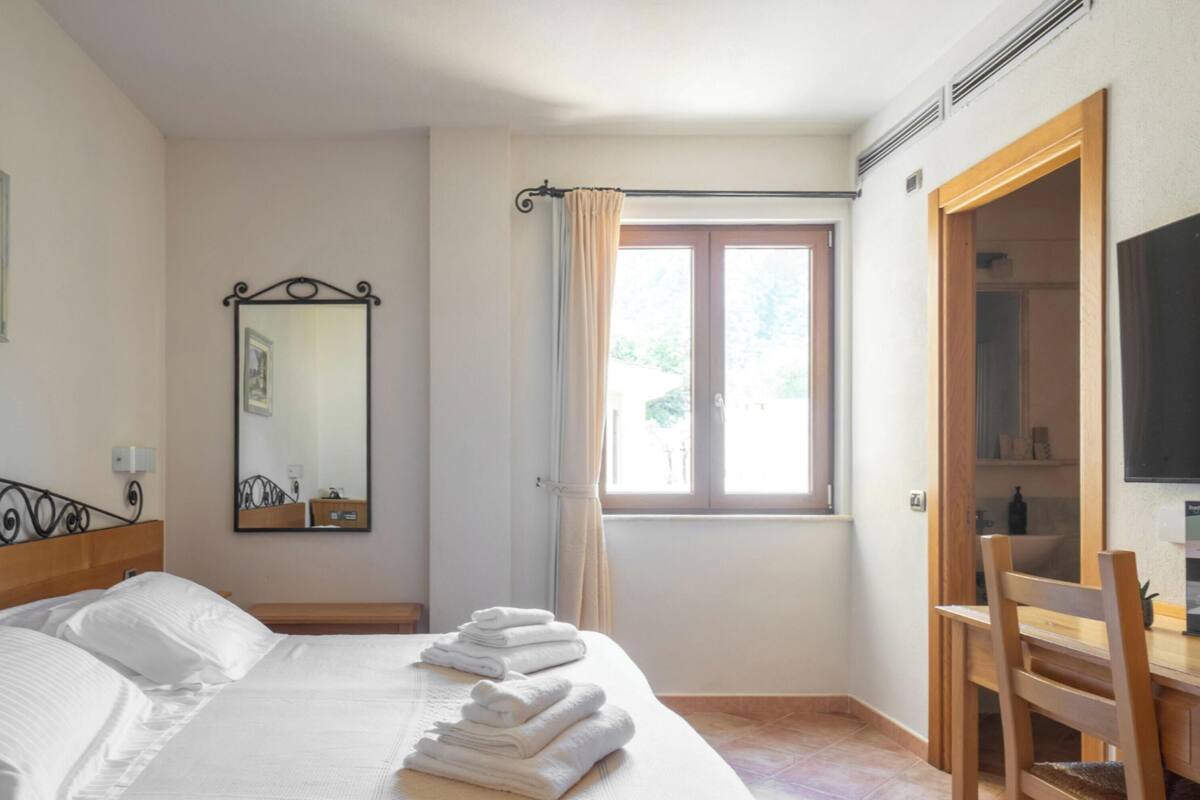 Una camera d'albergo con un letto, una scrivania, una sedia, uno specchio, una finestra con tende e una televisione.