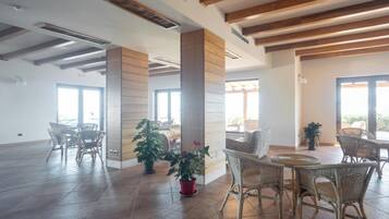 Ein geräumiges Zimmer mit hohen Decken, Holzbalken und großen Fenstern. Korbstühle und Tische sind zum Essen angeordnet.