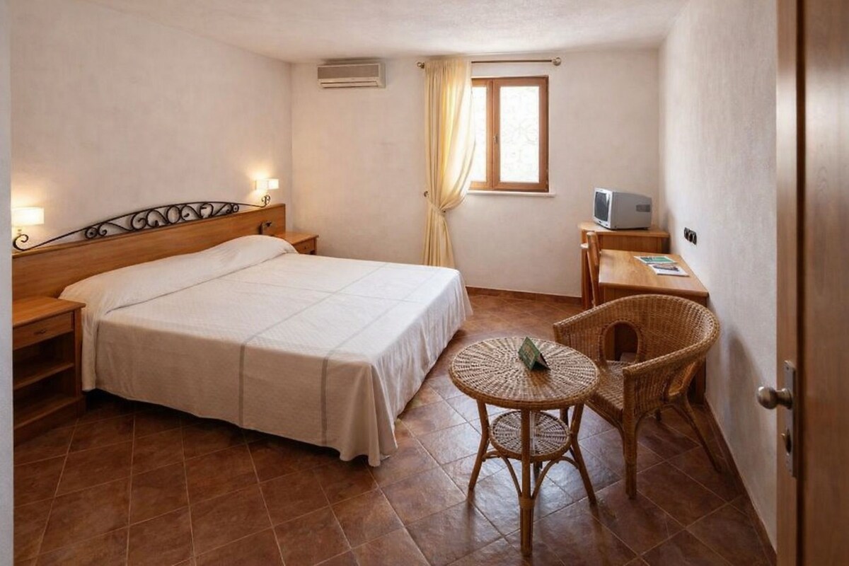 A bedroom with a bed, a small table, a chair, a television, and a window with curtains.