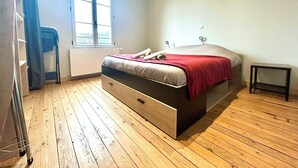 Een slaapkamer met een houten vloer, een bed met een rode deken, een raam en een klein nachtkastje.