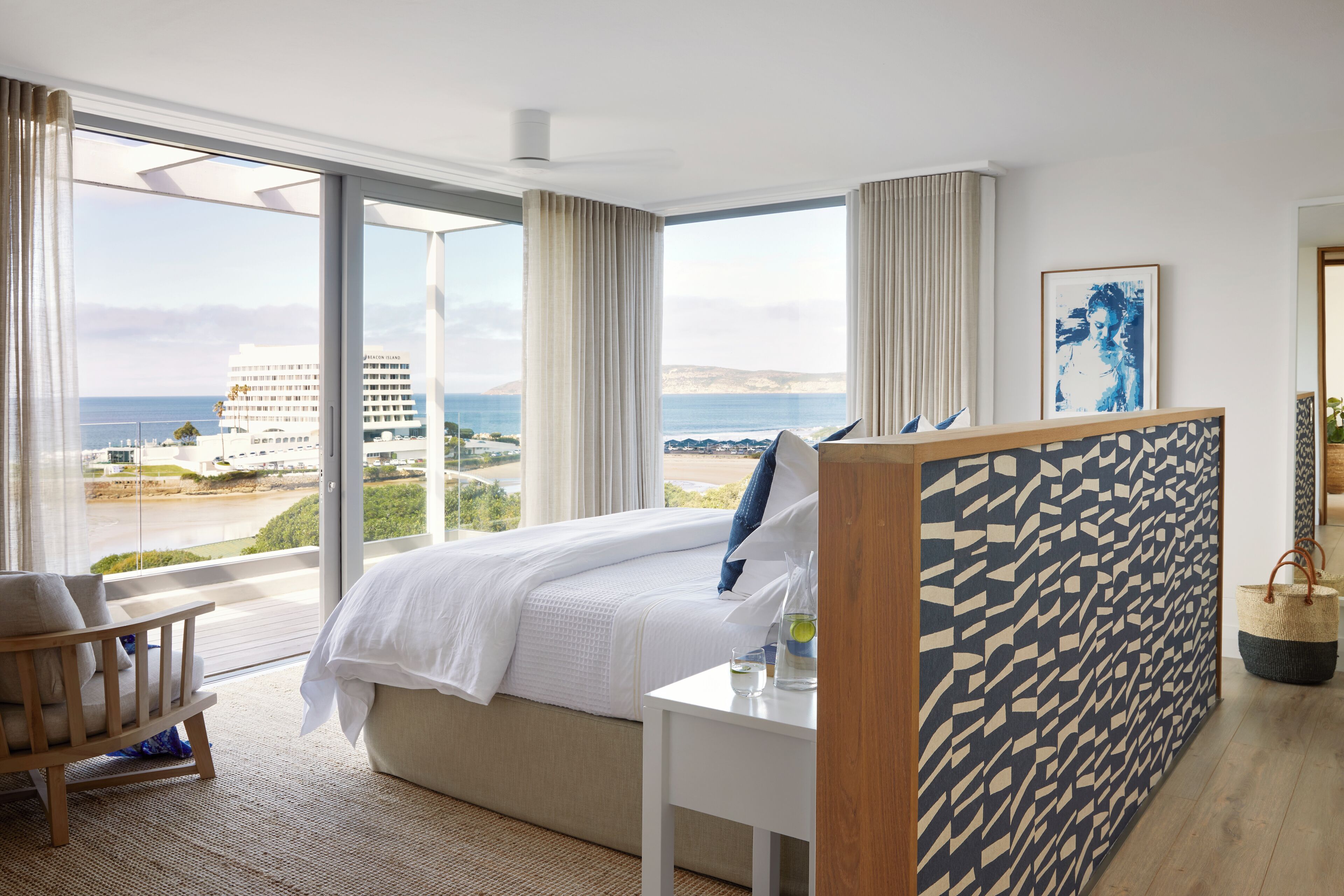 Un dormitorio con una ventana grande, una cama, una mesita de noche, una silla y vistas al mar y edificios.
