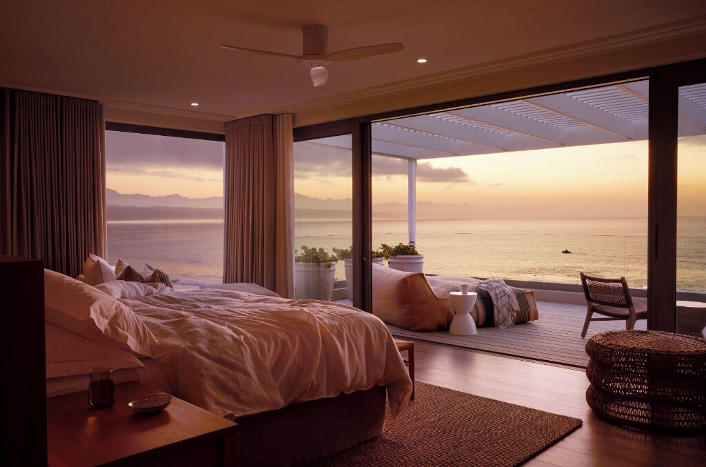 A bedroom with a large bed, a bedside table, a chair, and a view of the sea.