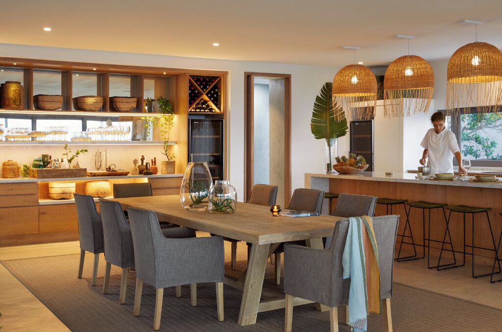 A modern kitchen with a wooden dining table, bar stools, and a person cooking.