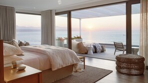 A bedroom with a large bed, a chair, a wicker ottoman, and a view of the sea. - Walkers Beach (Plettenberg Bay)