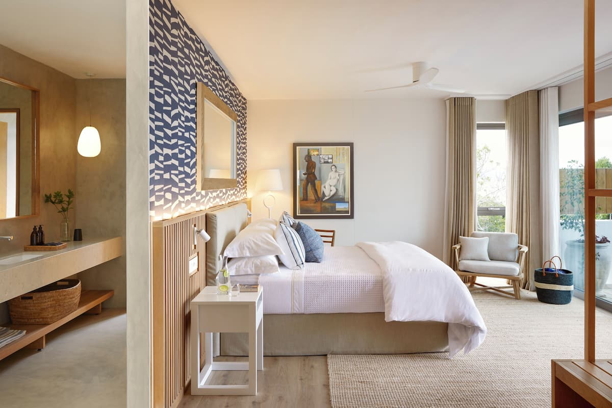 Una moderna camera d'hotel con un letto grande, un angolo relax con una poltrona, un tavolino con una lampada, uno specchio ampio e opere d'arte alle pareti.