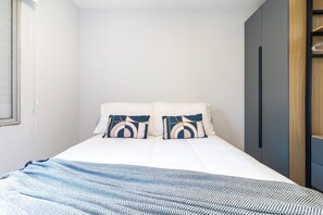 Cama bem arrumada com lençóis brancos e duas almofadas decorativas, um cobertor com padrão azul e branco, e um guarda-roupa de madeira à direita.