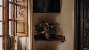 Una habitación con puerta de madera, televisor montado y estante con un arreglo floral.