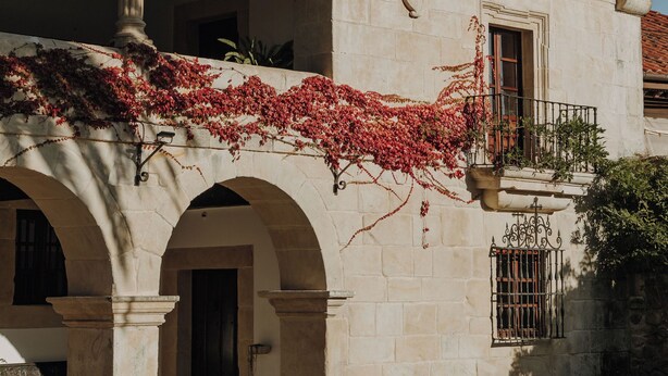 Un edificio histórico con fachada de piedra, entrada en arco y un balcón cubierto de enredaderas con flores rojas.