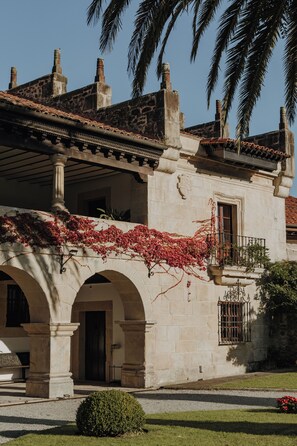 Un edificio histórico con fachada de piedra, entrada en arco y un balcón cubierto de enredaderas con flores rojas.