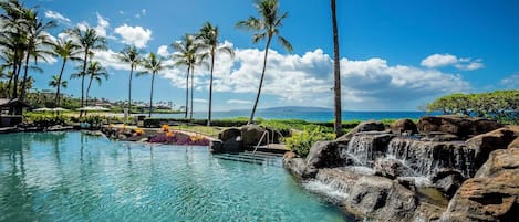 A clear pool with a waterfall feature, surrounded by palm trees and a view of the ocean.