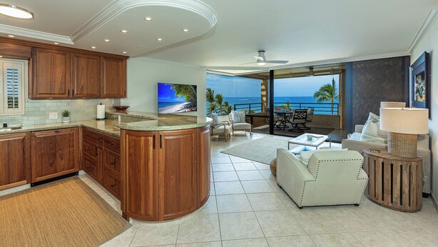 A modern kitchen with wooden cabinets, a central island, and a large TV. - Beachfront 2BR condo in Wailea w/ pool and hot tub (Wailea)