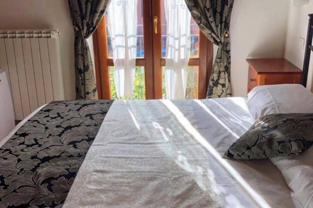 A bedroom with a bed, curtains, a radiator, and a view of greenery outside.