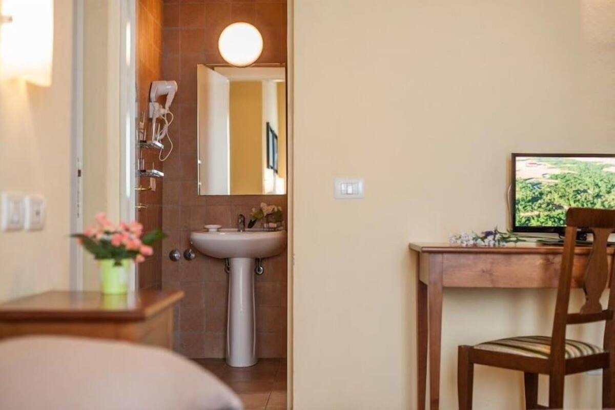 A bathroom with a sink, mirror, and hairdryer. There is a desk with a computer and a chair.