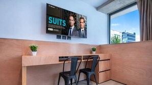 Una habitación de hotel moderna con un televisor grande en la pared, un escritorio de madera, dos sillas negras y una pequeña planta.