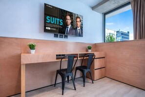 Una habitación de hotel moderna con un televisor grande en la pared, un escritorio de madera, dos sillas negras y una pequeña planta.
