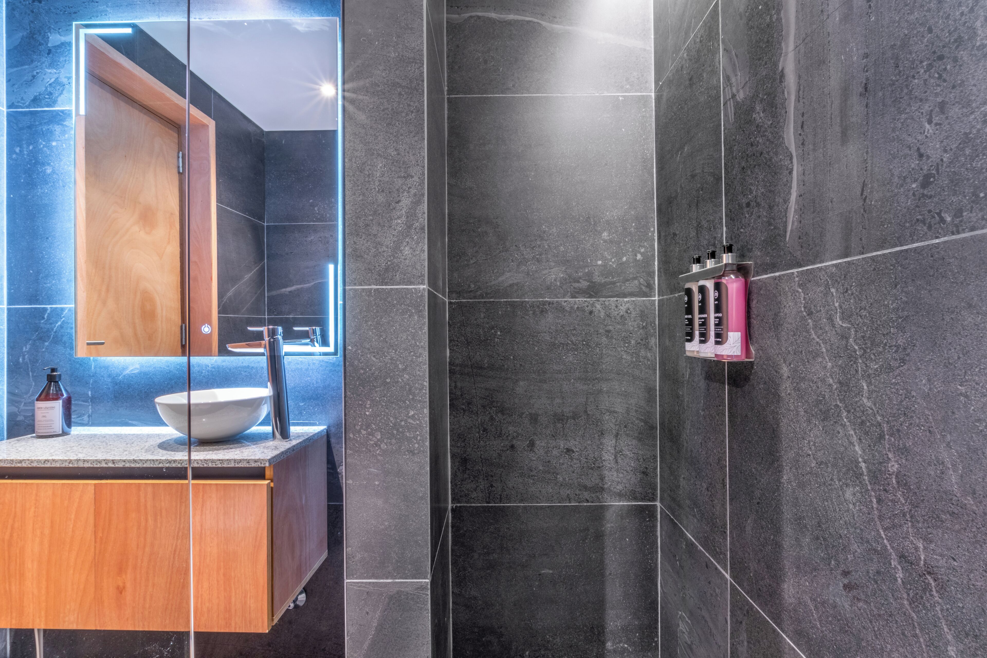 A modern bathroom with a wooden vanity, a large mirror, and a shower area with dark tiles.