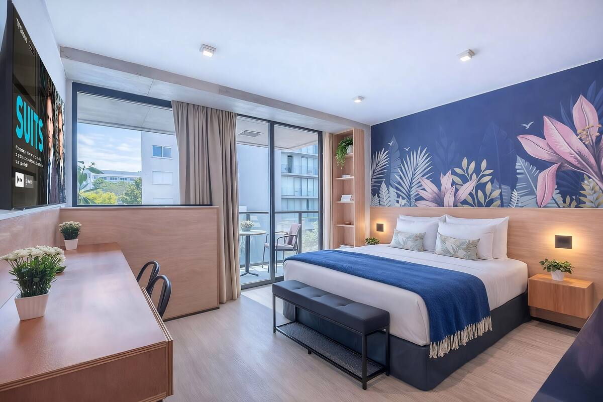 Une chambre d’hôtel moderne dotée d’un grand lit, d’un bureau, d’une banquette, d’une armoire, d’une porte-fenêtre coulissante donnant sur un balcon et d’une fresque murale.