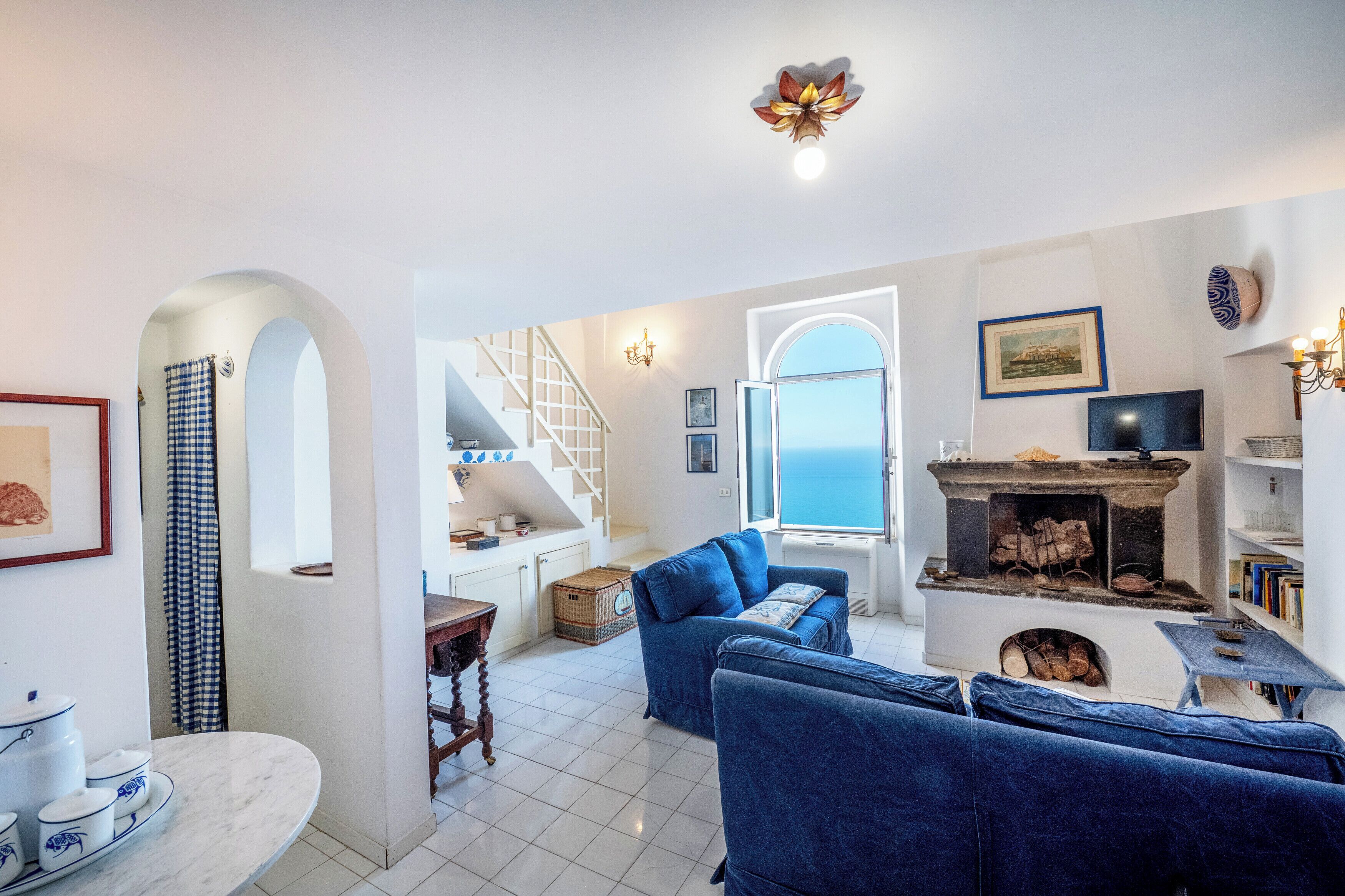 A living room with a fireplace, blue sofas, a staircase, and a view of the sea.
