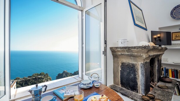 A breakfast table with a view of the sea, a stone fireplace, and a bookshelf. - Casa Capo di Conca by Wonder Tour (Conca dei Marini)