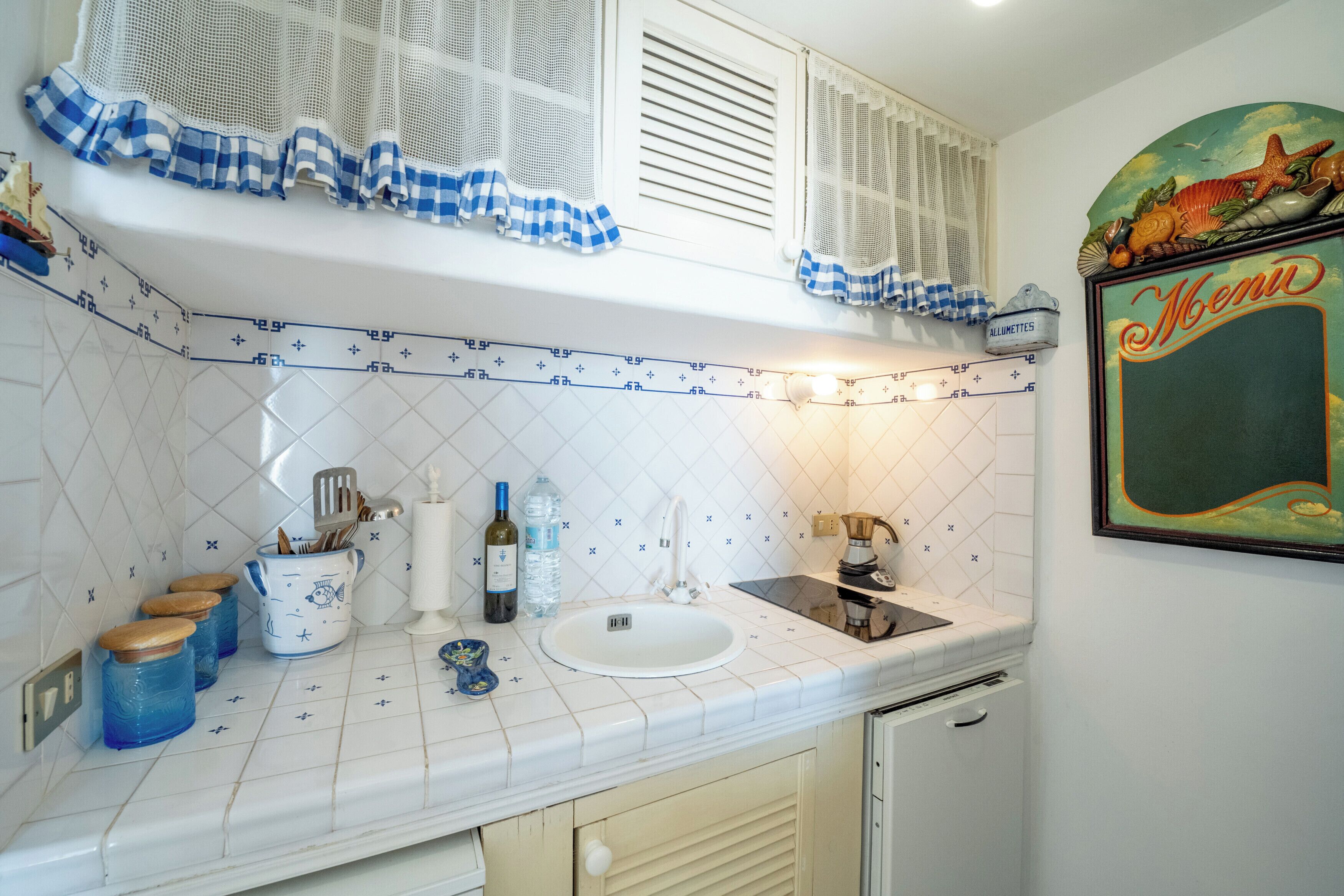 A kitchen with a white tiled backsplash, a white sink, and a window with blue and white checkered curtains.