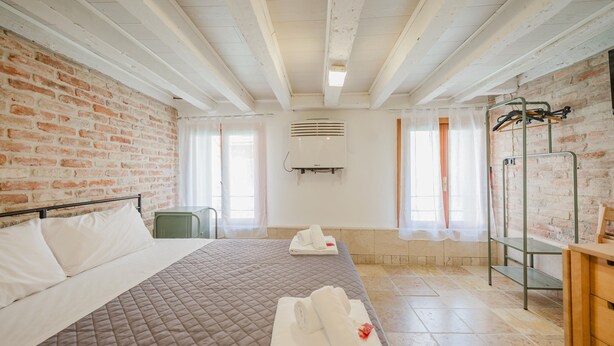 A hotel room with a brick wall, a bed with a quilted bedspread, two rolled towels, a green chair, a wooden bench, and a metal rack with clothes. - Balastro Design Rooms (Venice)