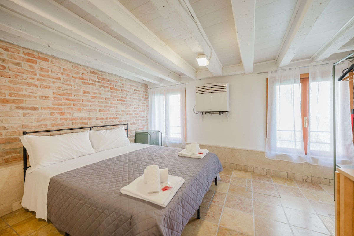 Quarto de hotel com parede de tijolos, uma cama grande com colcha acolchoada, duas mesas de cabeceira com toalhas, uma janela com cortinas e um ar condicionado fixado na parede.