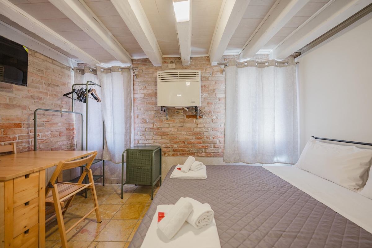 Quarto de hotel com parede de tijolos, cama com colcha acolchoada, escrivaninha de madeira com cadeira e radiador.