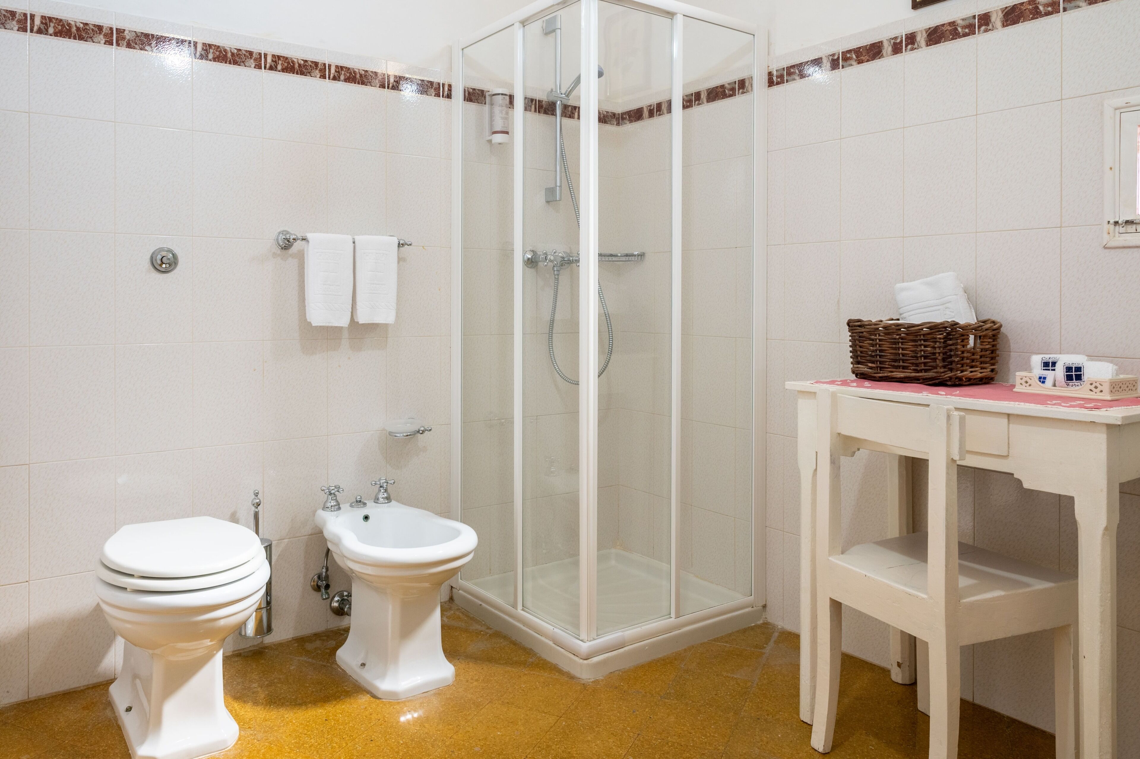 A bathroom with a toilet, bidet, shower, and a small table with a basket.