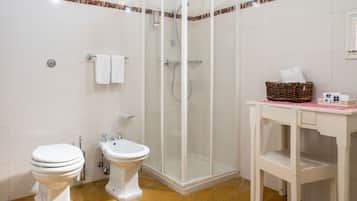 A bathroom with a toilet, bidet, shower, and a small table with a basket.