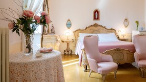 Un dormitorio con una cama, dos sillas, un tocador con espejo, una mesa con un jarrón de flores y un icono religioso colgado en la pared.