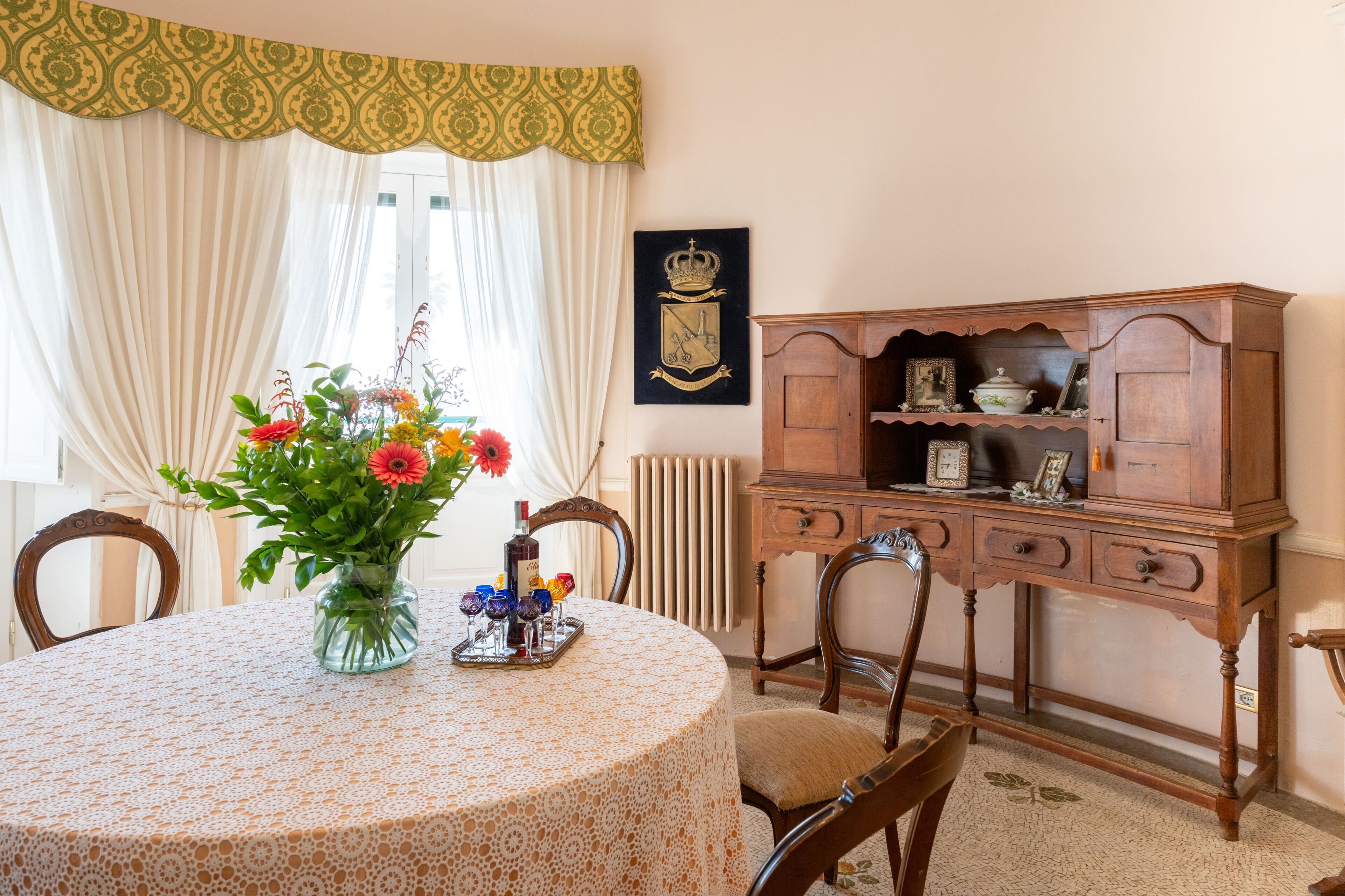 A round table with a floral tablecloth, a vase with flowers, and a tray with glasses and a bottle.
