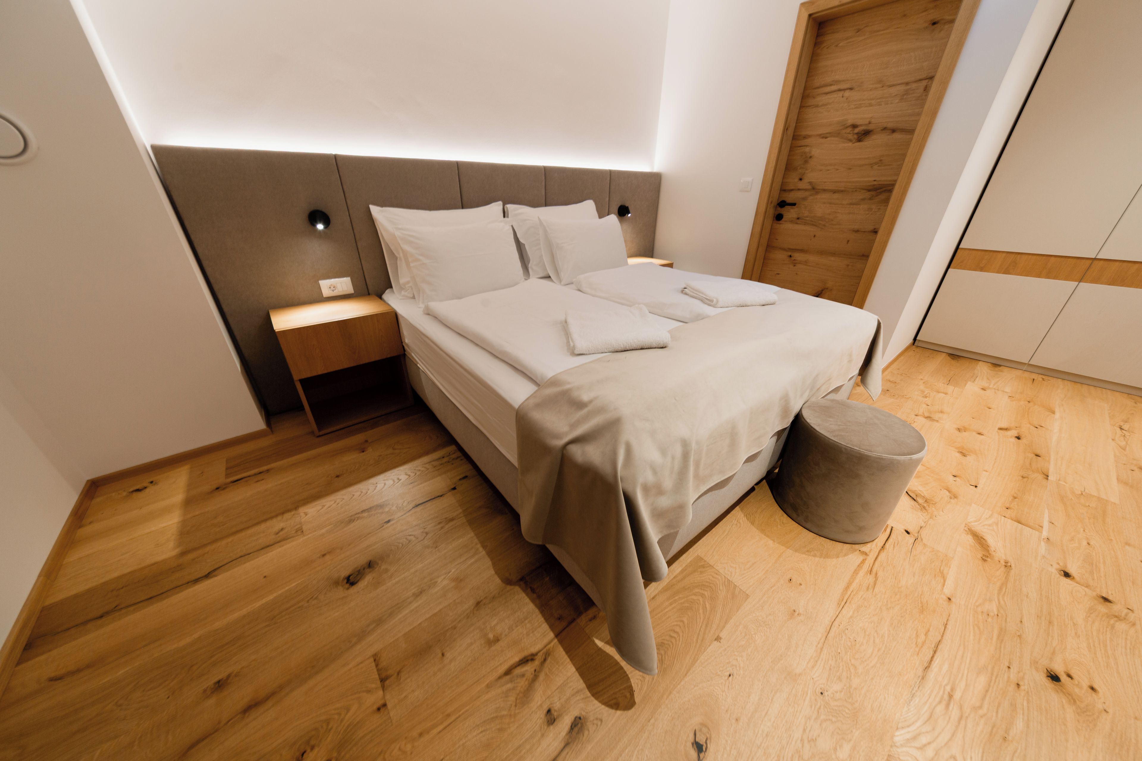 A modern bedroom with a wooden floor, a bed with white linens, a nightstand, and a wooden door.