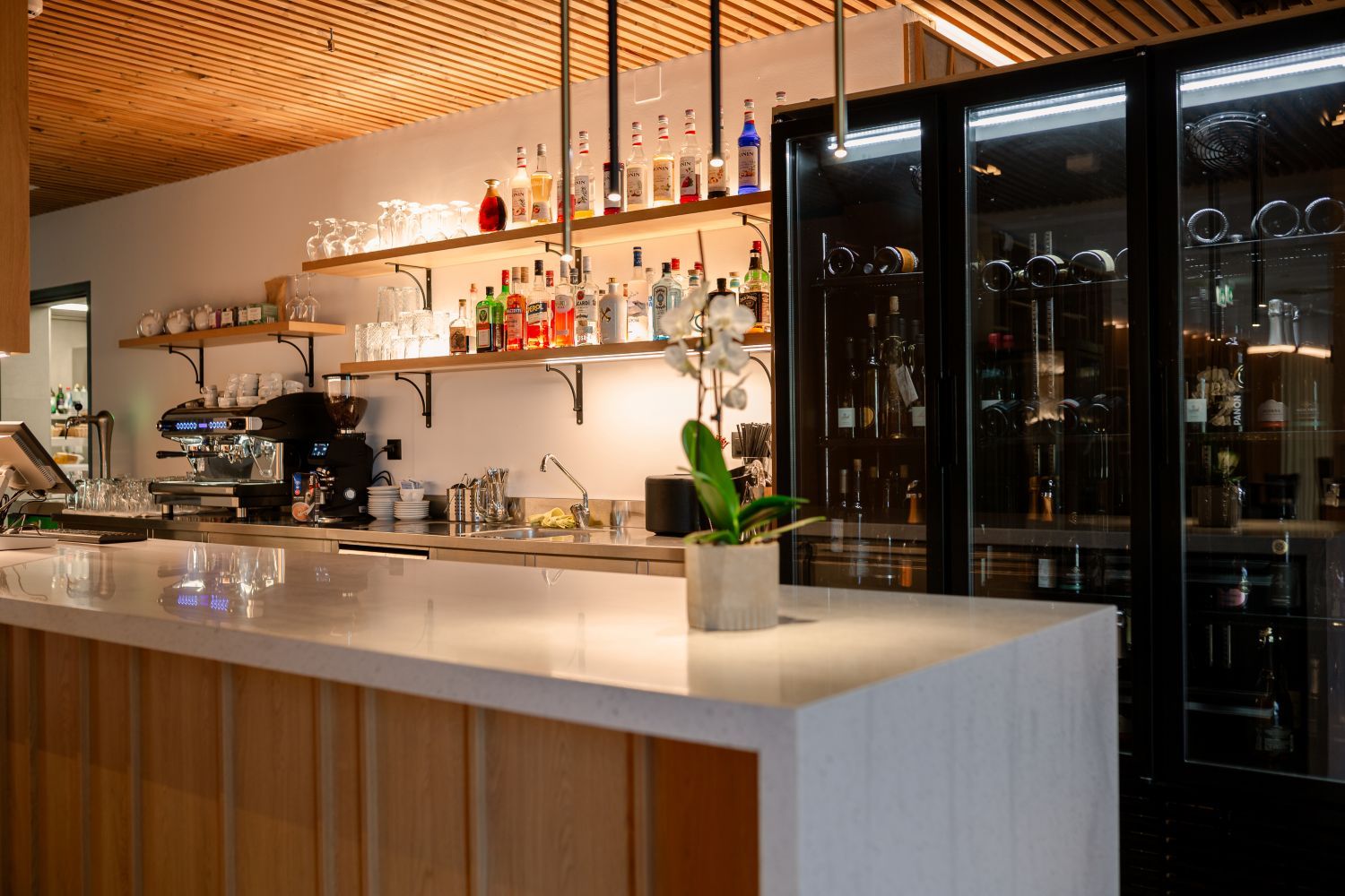 A bar area with a countertop, shelves with bottles, a coffee machine, and a refrigerator.