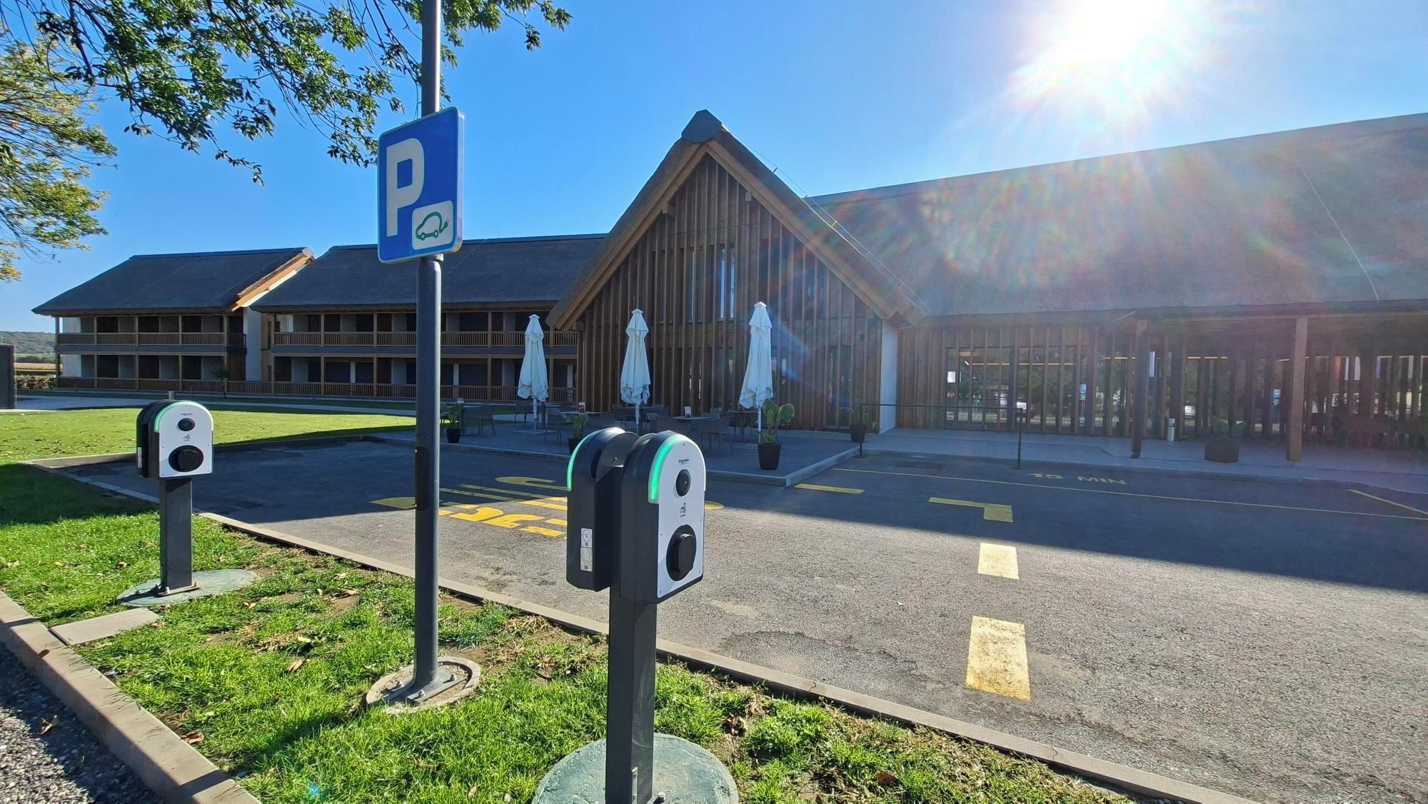 Un espace de stationnement équipé de bornes de recharge pour véhicules électriques, un bâtiment à l’architecture moderne et un espace extérieur aménagé avec des sièges.