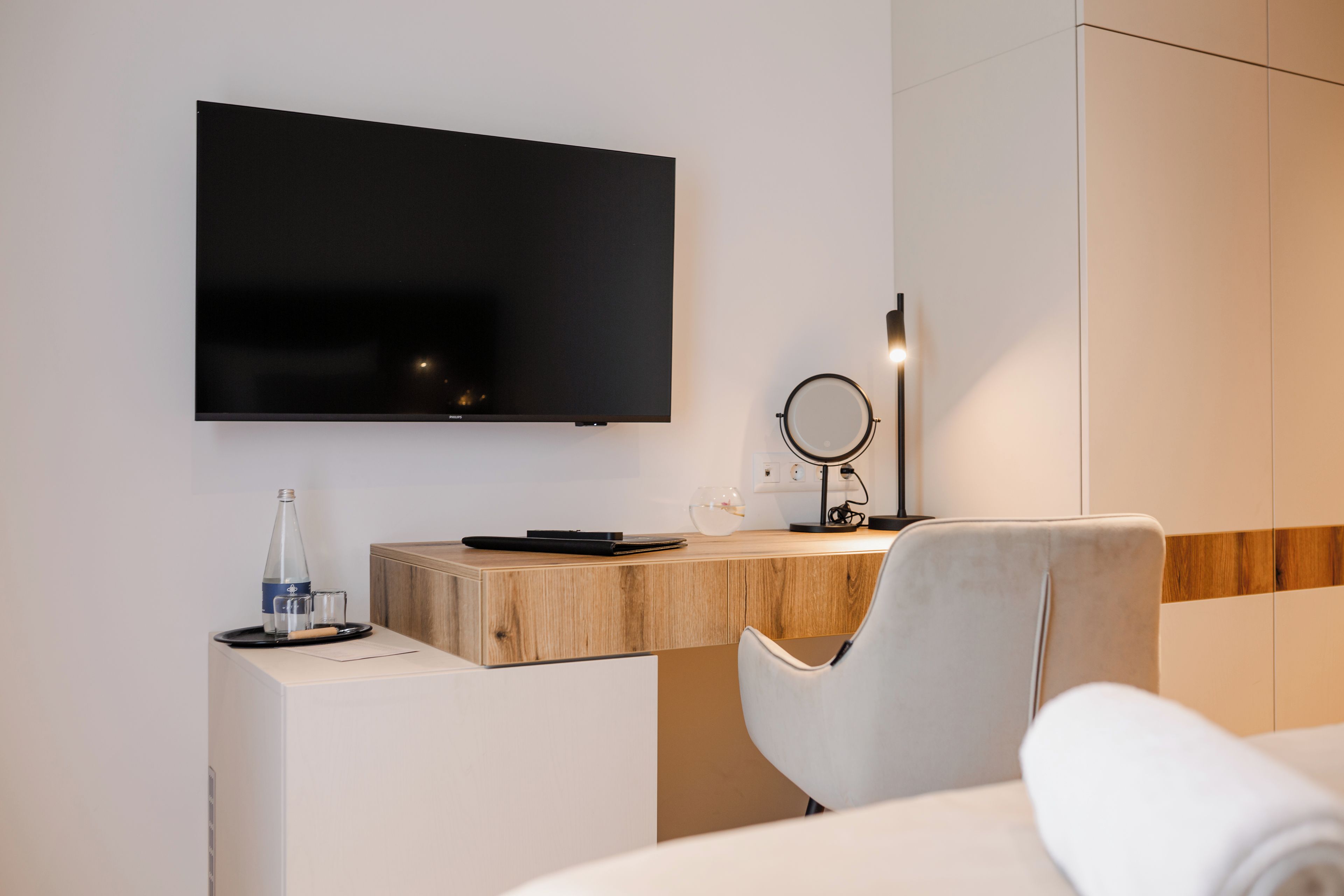 Une chambre d’hôtel moderne équipée d’un téléviseur à écran plat fixé au mur, d’un bureau en bois avec un ordinateur portable, d’un miroir rond et d’une chaise blanche.