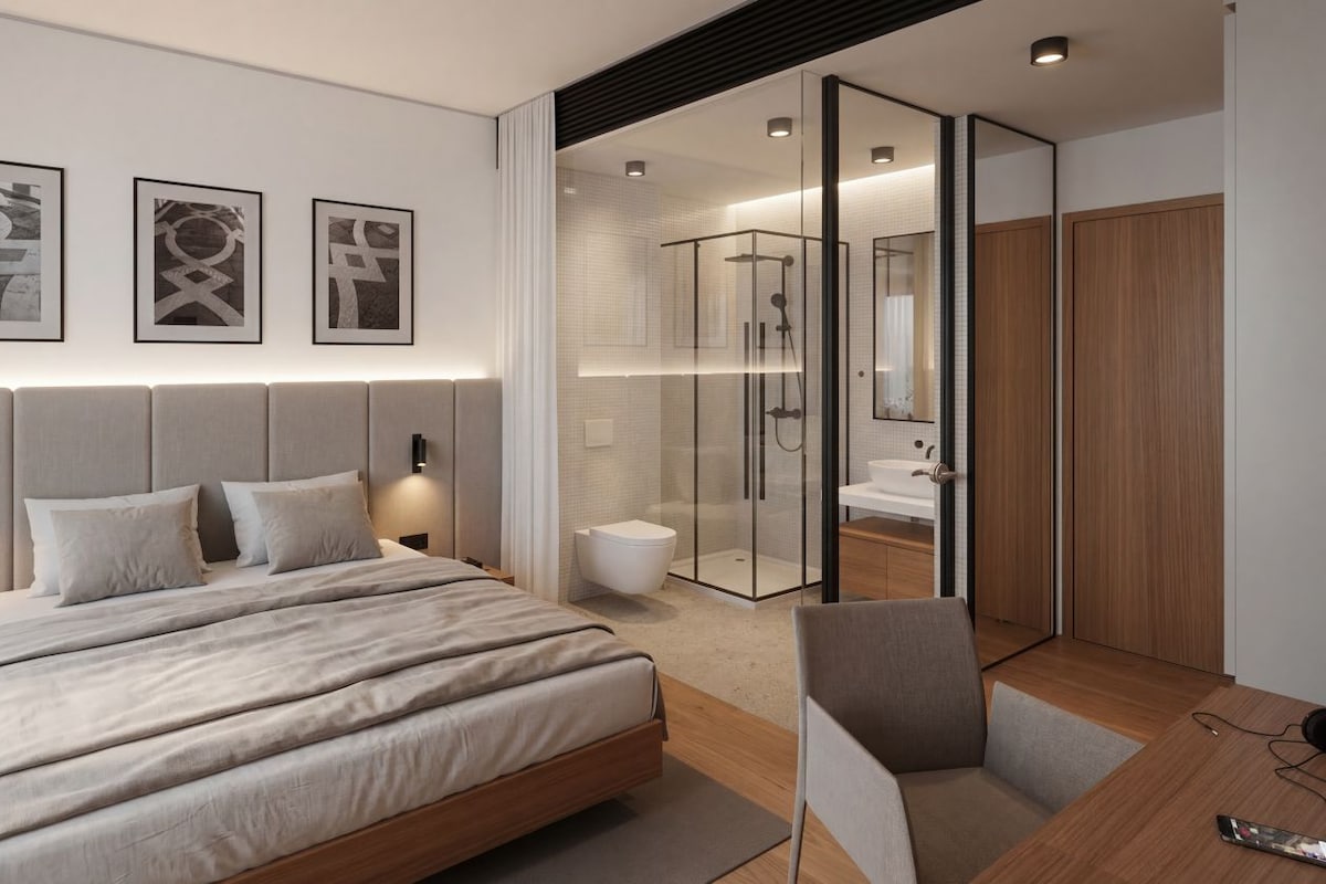 Un dormitorio moderno con una cama grande, mesitas de noche, una silla, ducha con cerramiento de vidrio y un armario de madera.
