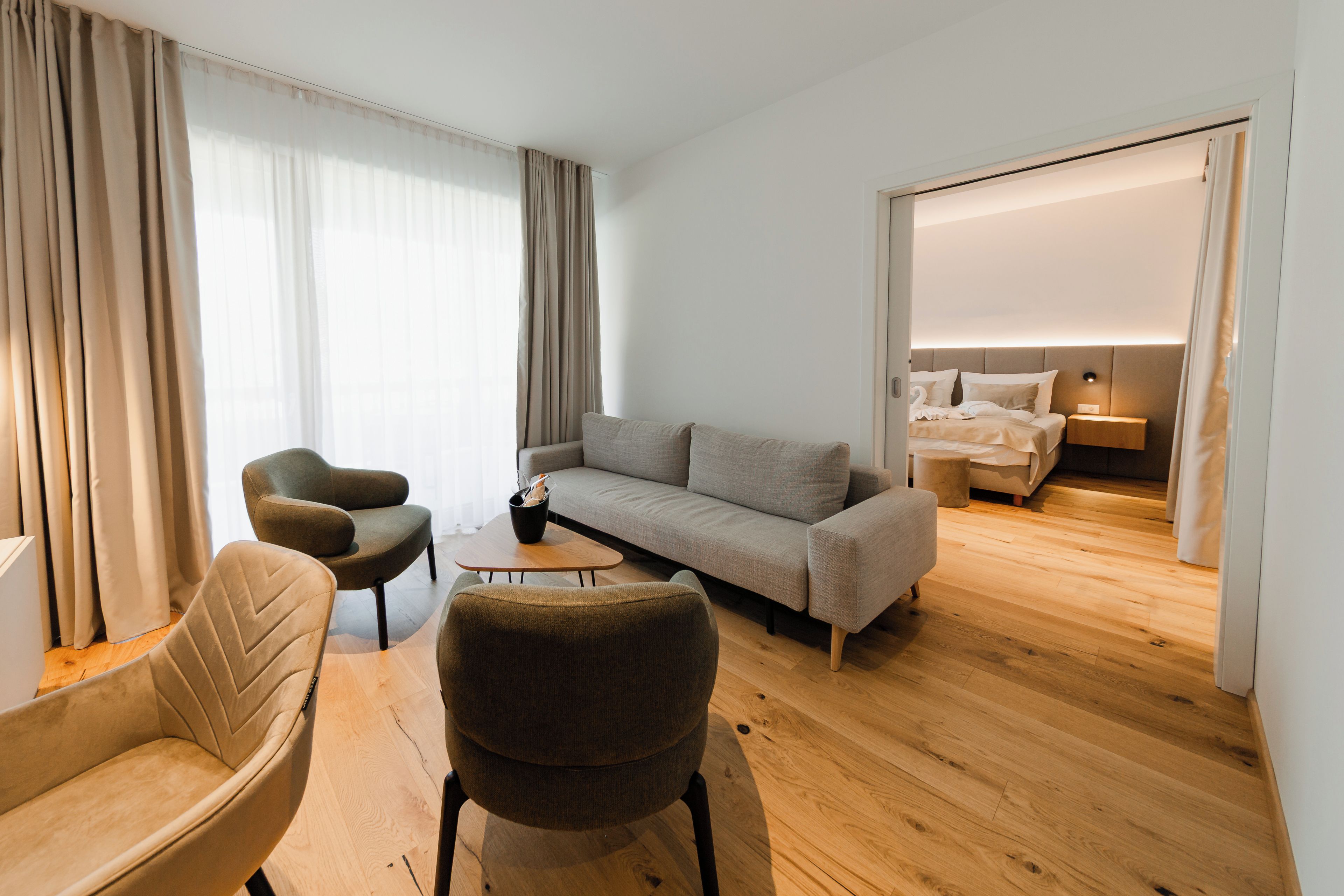 A modern hotel room with a sofa, armchairs, a small table, and a bed with a nightstand.