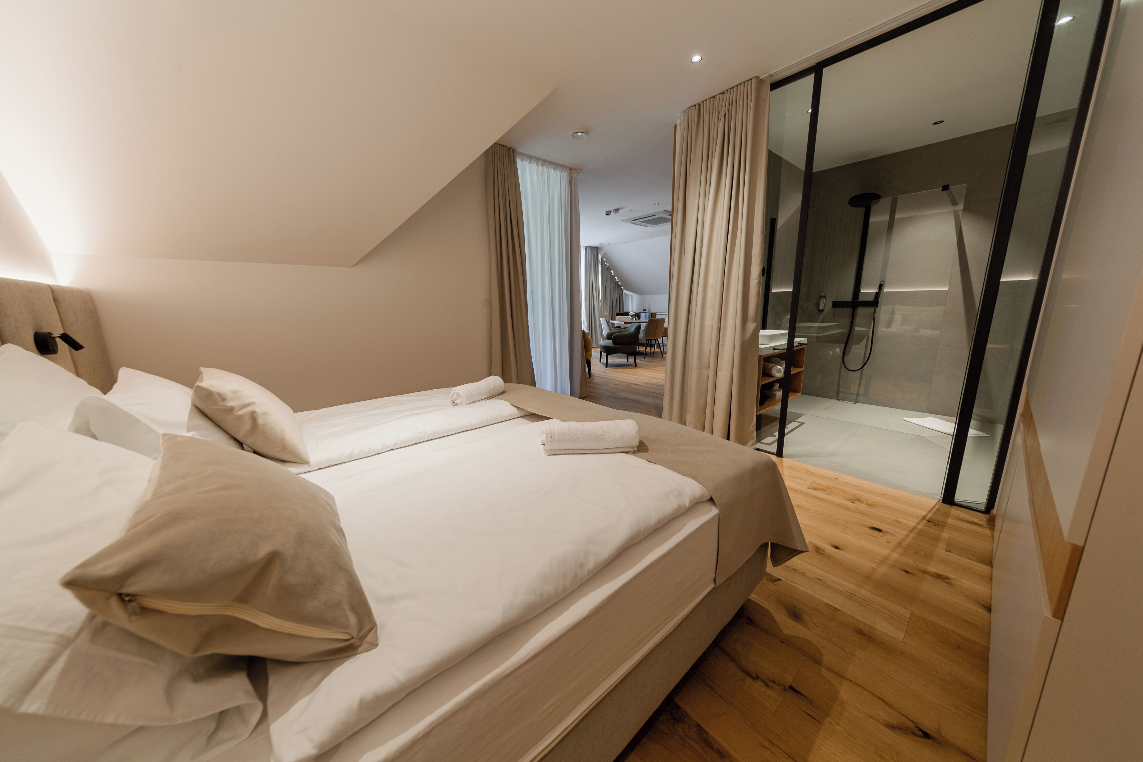 A modern hotel room with a large bed, a glass shower enclosure, and a view of the dining area.