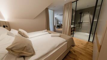 A modern hotel room with a large bed, a glass shower enclosure, and a view of the dining area.