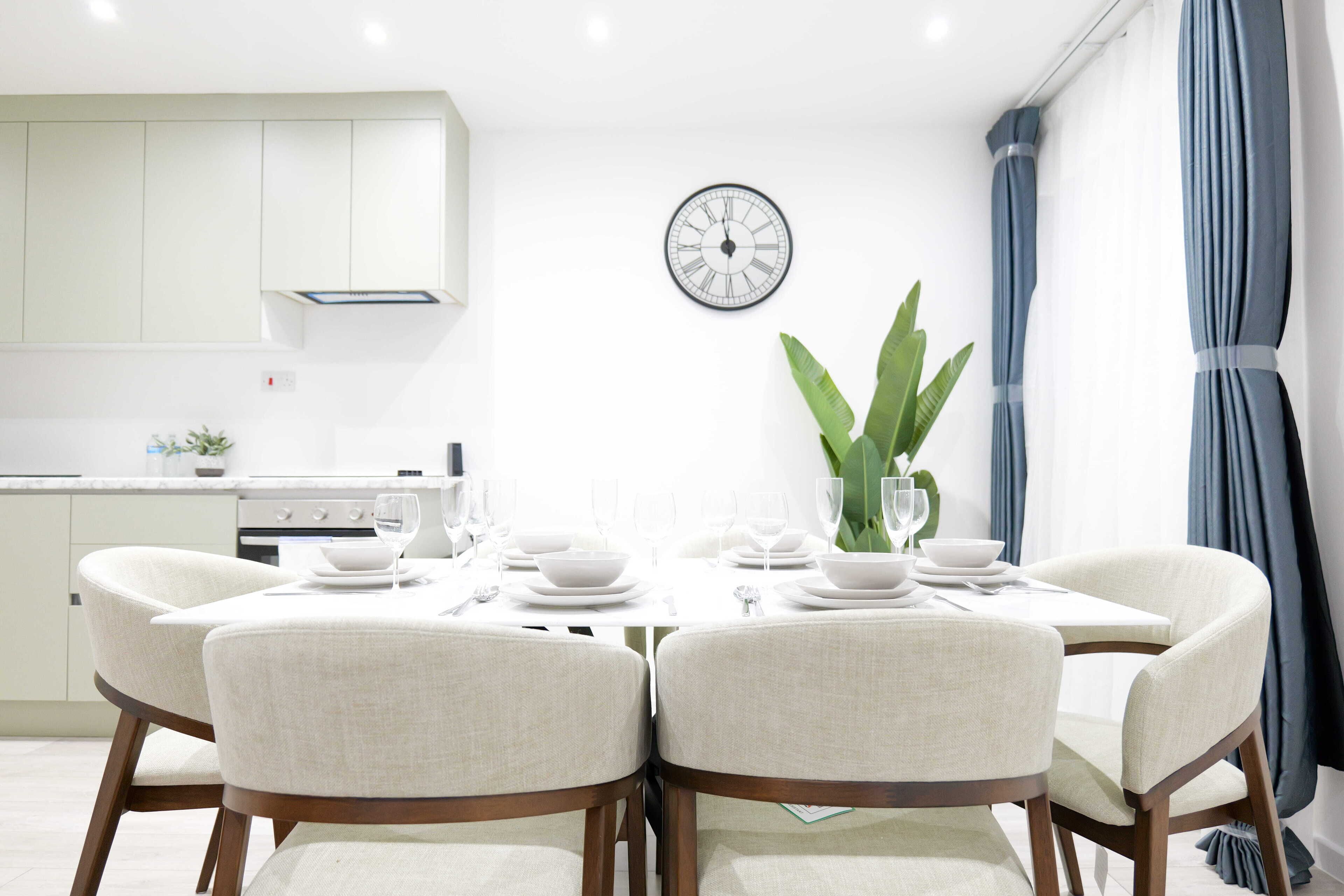 Uma sala de jantar moderna com uma mesa redonda posta para quatro pessoas, cadeiras brancas, um relógio de parede e um vaso com planta.