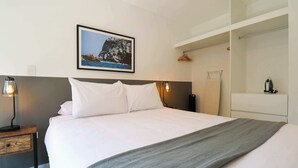 Una cama bien hecha con sábanas blancas y una manta gris, una mesita de noche con lámpara, un cuadro con un paisaje costero, y un vestidor con perchero.