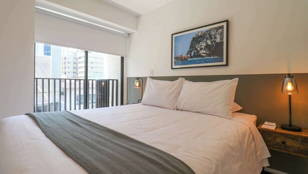 Habitación de hotel con una cama grande, un balcón con vista a edificios, un cuadro en la pared y una mesita de noche con una lámpara.