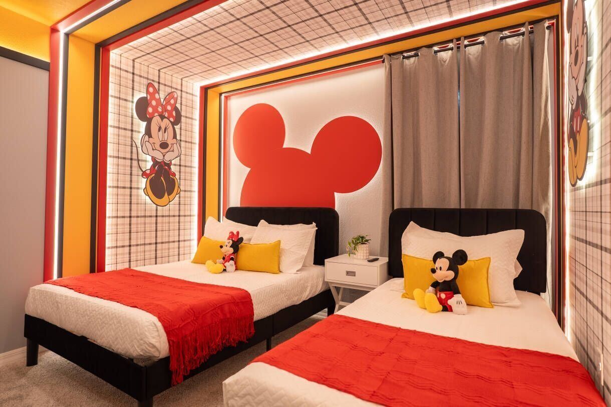 A hotel room with two beds, each adorned with Mickey Mouse-themed pillows and a red blanket.