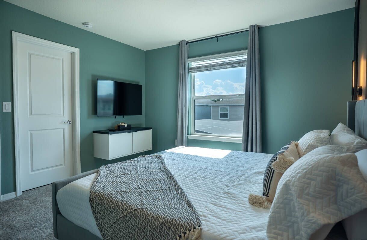Une chambre à coucher avec un lit, un téléviseur fixé au mur, une fenêtre avec des rideaux et une vue sur l’extérieur.