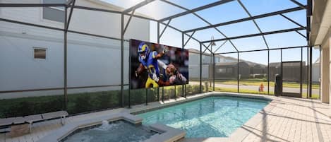 Zona de piscina con jacuzzi, pantalla grande mostrando deportes y porche cubierto.